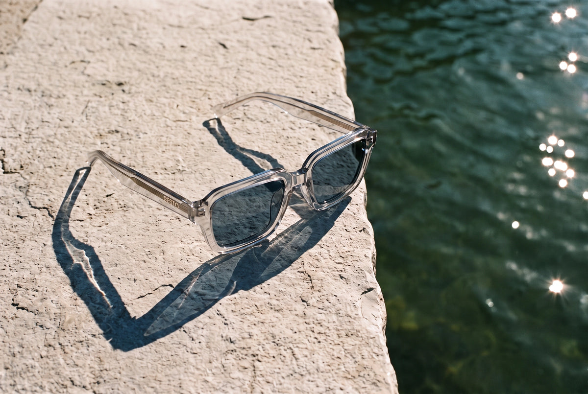 SESTO. SORA Sonnenbrille mit grau-blauen Gläsern auf Stein am Wasser, Spiegelungen auf der Oberfläche